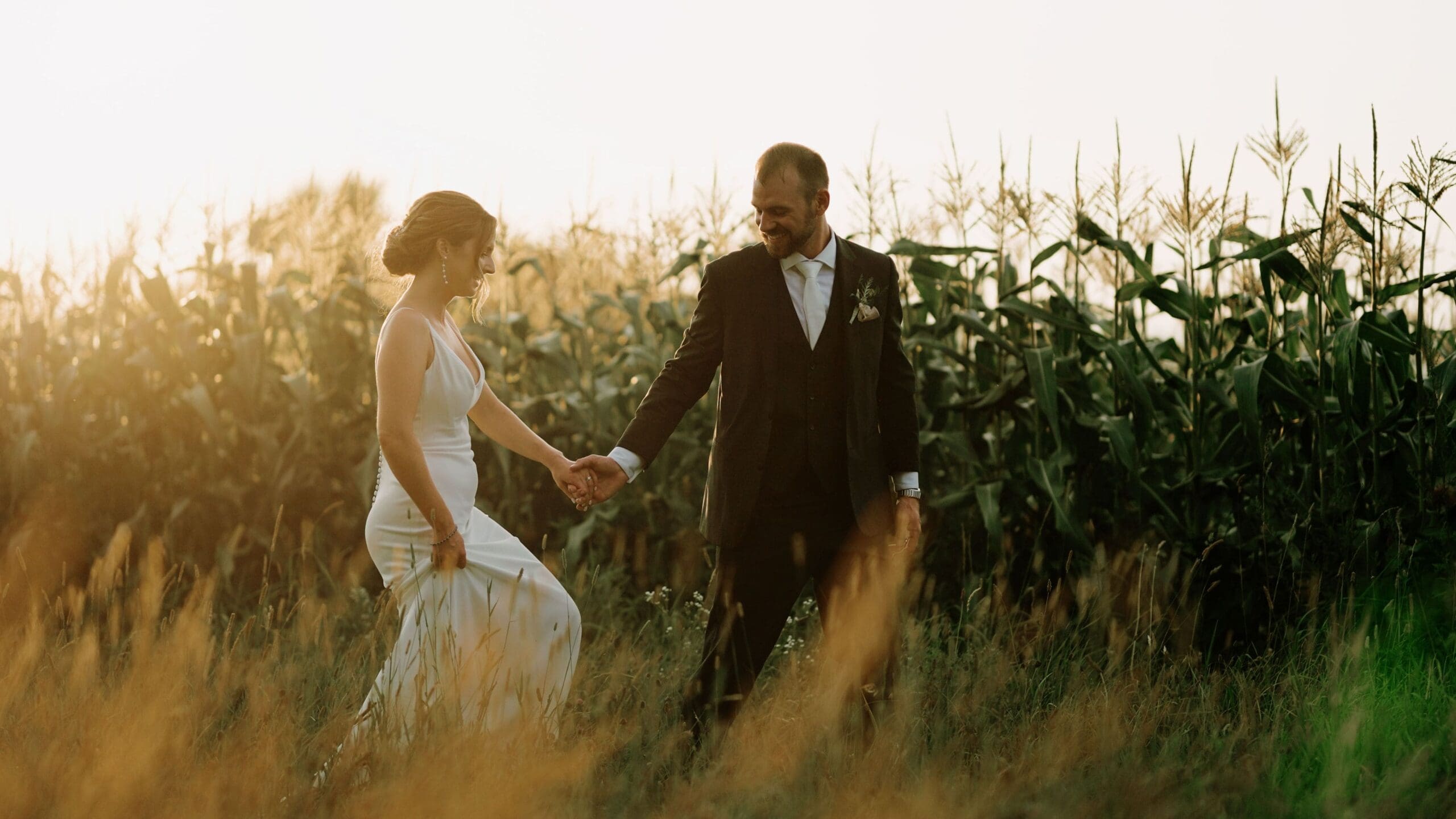 Bride And Groom Walking Through Fields With The Sun Setting
