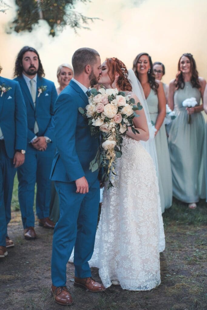 Couple Kissing, With Wedding Party Behind Them In Outdoor Reception
